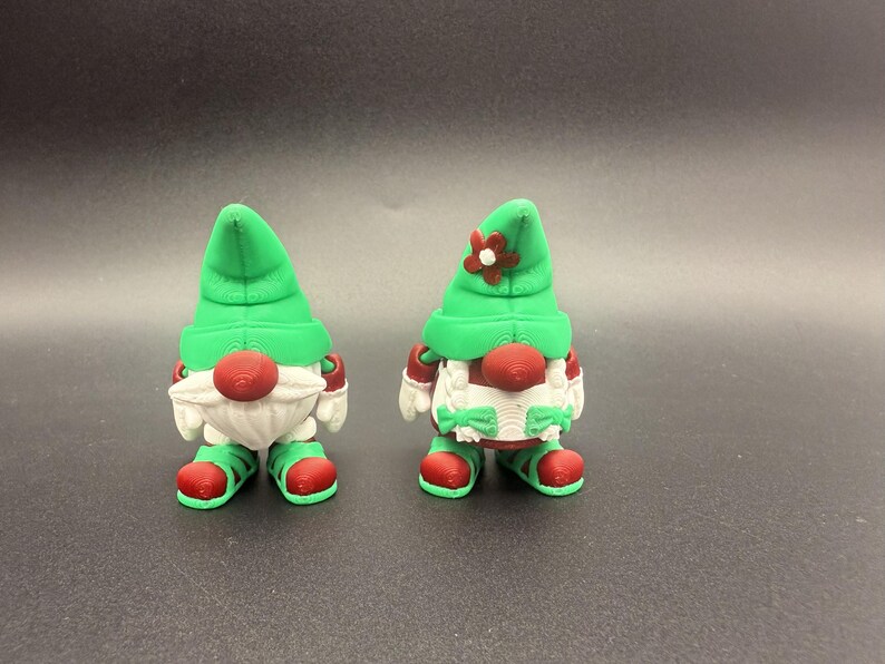 Puede incluir: Dos figuras de gnomos caprichosos. Cada gnomo tiene un gorro puntiagudo verde, una nariz roja y una barba blanca. Llevan zapatos y mitones rojos. Un gnomo tiene una flor en su sombrero. Para decoraci&oacute;n navide&ntilde;a o de jard&iacute;n.