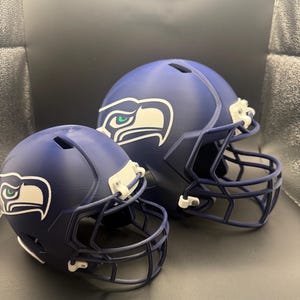 Peut inclure: Deux casques de football bleu marine, un grand et un petit, sont présentés. Chaque casque arbore un logo d'oiseau blanc et vert. Les casques ont des masques faciaux bleu foncé et des clips blancs.