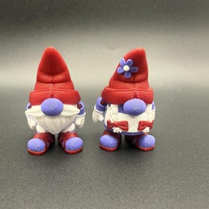 Puede incluir: Dos figuras de gnomos caprichosos. Cada gnomo tiene un sombrero rojo, una barba blanca y detalles morados en la nariz, las manos y los pies. Un gnomo tiene una flor en su sombrero. Los gnomos miden aproximadamente 7,6 cm de altura.