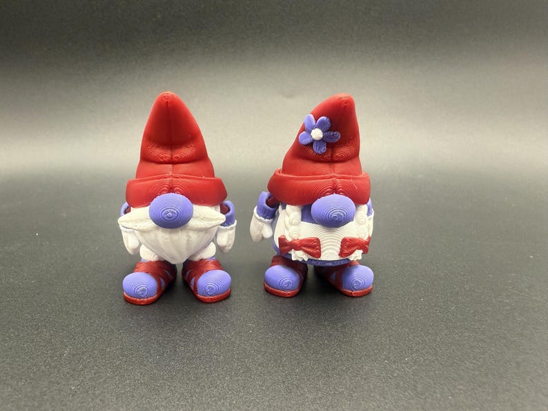 Puede incluir: Dos figuras de gnomos caprichosos. Cada gnomo tiene un sombrero rojo, una barba blanca y detalles morados en la nariz, las manos y los pies. Un gnomo tiene una flor en su sombrero. Los gnomos miden aproximadamente 7,6 cm de altura.