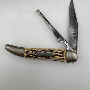 May include: A folding pocket knife with a cream-colored handle featuring a dark brown textured pattern and a metal shield inlay stamped with the text FISH KNIFE. The knife has two open metal blades: one standard blade and one serrated scaler.