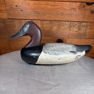 May include: A carved wooden duck decoy featuring a dark brown head, a black chest, and a body with light gray and white painted details. The surface shows signs of wear and texture, with a pointed black beak and a streamlined, horizontal posture.