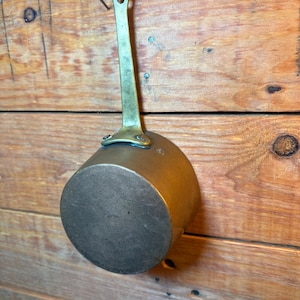 May include: A small, antique copper saucepan with a long brass handle. The round pot is suspended from a wooden wall by its handle. The copper has a dark, aged patina, and the handle is attached with rivets.