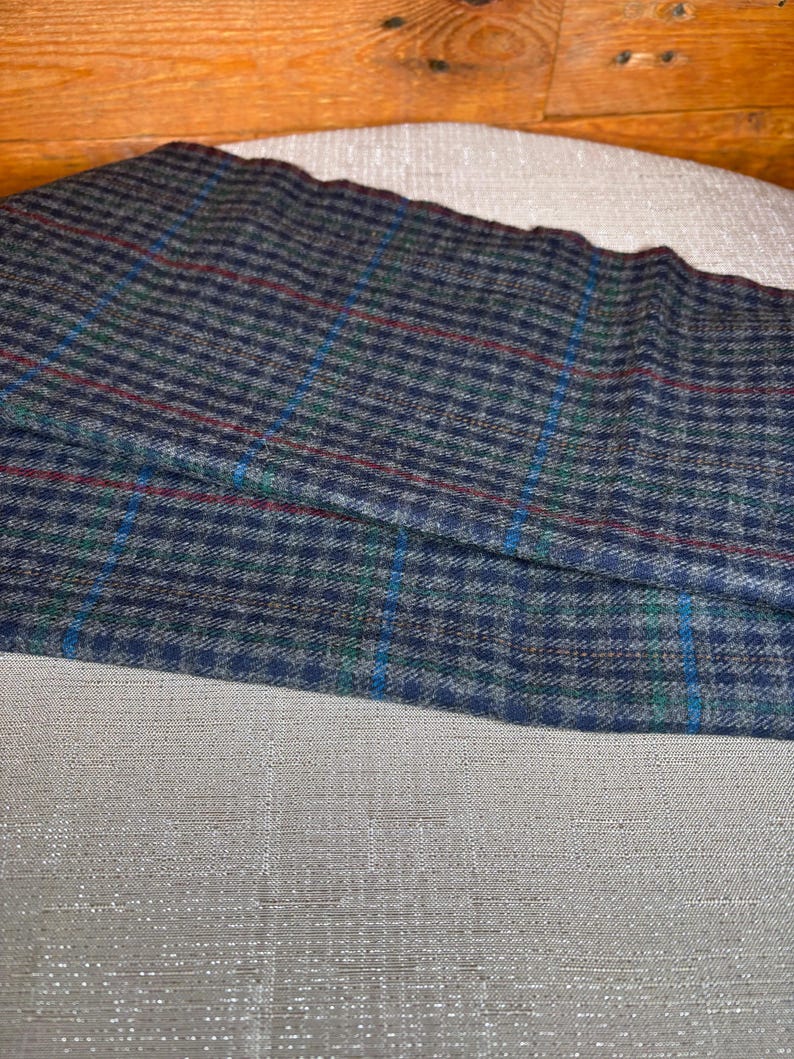 May include: A folded piece of fabric with a blue, green, and red plaid pattern. The fabric is a dark blue with a small checkered design. The plaid includes thin lines of red, green, and blue. The fabric is resting on a light grey surface.