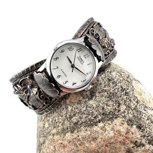 May include: A silver-toned wristwatch with a white face, black numerals, and the text Timex Indiglo 30 Meters. The watch is integrated into a wide, ornate silver cuff bracelet featuring detailed floral and leaf metalwork engravings.