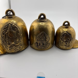 May include: Three antique brass bells of varying sizes. Each bell features a loop for hanging and a detailed embossed design of a seated figure. The bells have a warm, golden hue and a slightly aged appearance.