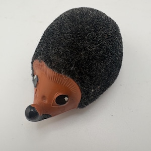 May include: A hedgehog figurine featuring a smooth, light brown terracotta face with a black nose and black eyes. The body is covered in a dense, dark charcoal-colored textured material that mimics the appearance of quills or fur.
