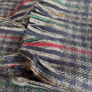 May include: Close-up of a woven textile with a fringed edge. The fabric features a navy blue, grey, and white plaid pattern with red, green, and gold accent stripes. The fringed edge adds texture and visual interest to the textile.
