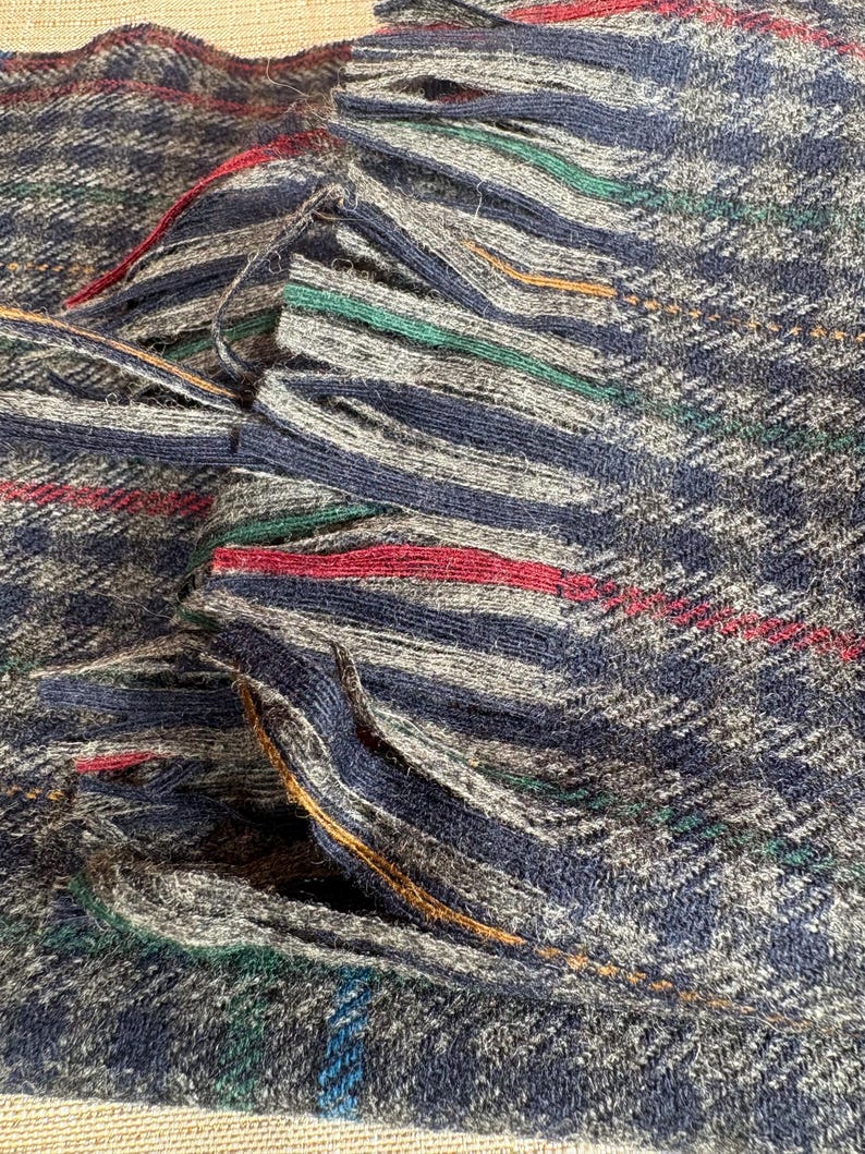 May include: Close-up of a woven textile with a fringed edge. The fabric features a navy blue, grey, and white plaid pattern with red, green, and gold accent stripes. The fringed edge adds texture and visual interest to the textile.