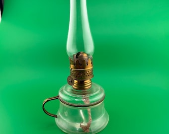 May include: A clear glass oil lamp with a brass-colored metal base and a long, clear glass chimney. The lamp has a small handle and a wick visible inside. The lamp is set against a bright green background.