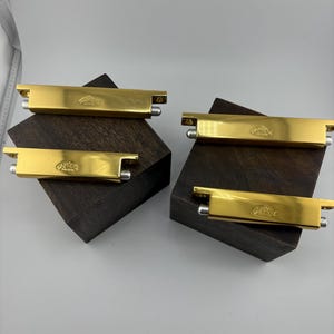 May include: Four gold-colored metal clips with a rectangular shape and rounded ends. Each clip is engraved with a small emblem. The clips are displayed on dark brown wooden blocks against a neutral background.