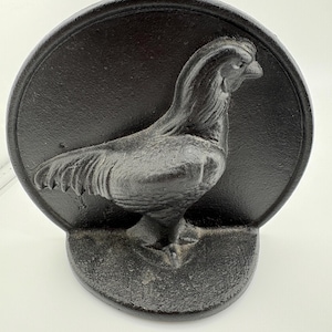May include: A black cast iron rooster doorstop. The rooster is in profile, with detailed feathers and a prominent comb. The doorstop is circular with a semi-circular base, designed to stand upright.