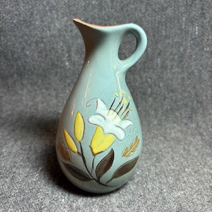 May include: A small, turquoise ceramic pitcher with a handle and a spout. The pitcher is decorated with a white and yellow floral design with brown leaves. The rim of the pitcher is a darker brown color.