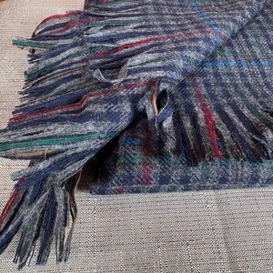 May include: A folded scarf with a plaid pattern in blue, grey, red, and green. The scarf has fringed edges. The fabric appears to be a soft, woven material. The scarf is folded over on itself, suggesting it is ready to wear.