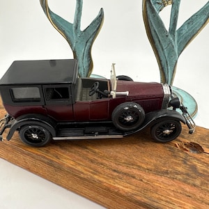 May include: A miniature vintage-style automobile model featuring a deep maroon body, a black roof, and black fenders. The car has black spoked wheels, a spare tire mounted on the side, and a visible steering wheel inside the open-top cabin area.
