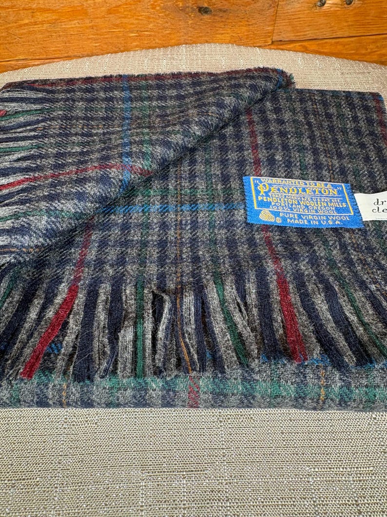 May include: A navy, grey, red, and green plaid wool scarf with a fringed edge. The scarf has a blue tag that reads "Pendleton" and "Pure Virgin Wool Made in U.S.A."
