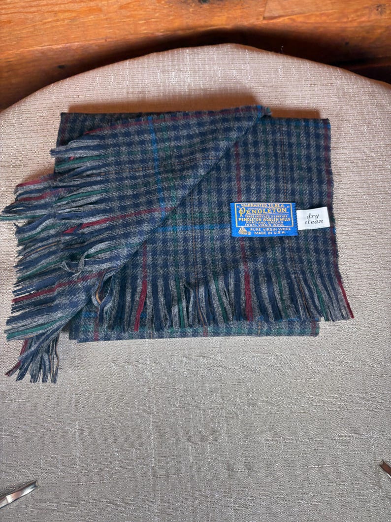 May include: A folded blue and grey checked scarf with fringed edges. The scarf features red and green accents and a blue label that reads "Pendleton". A small white tag reads "dry clean".