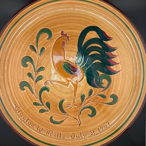 May include: A decorative ceramic plate featuring a rooster design. The plate has a tan background with a brown rim and green accents. The rooster is orange, yellow, and green. The plate is inscribed with "Clayton W. Mills, July 31, 1967."
