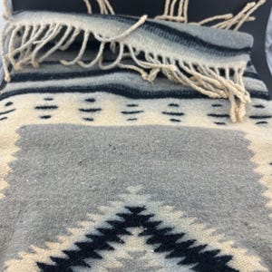 May include: A folded gray, cream, and black woven blanket with a geometric pattern. The blanket has a fringe along the edge. The design features diamond and horizontal stripe patterns. The blanket is made of a soft, textured material.