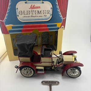 May include: A vintage Schuco Oldtimer Mercedes Simplex anno 1902 toy car with a wind-up key. The car is cream and burgundy with black accents and a black convertible top. The box is blue and gold with the Schuco logo.