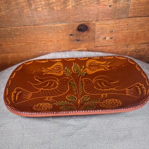 May include: A rectangular, brown ceramic serving dish with a decorative bird and floral design. The dish has a raised, scalloped edge and a salmon-colored rim. The birds are yellow and the leaves are green.
