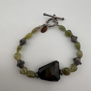 May include: A bracelet featuring a large, faceted dark brown stone centerpiece, accented by smaller, irregular light green stones and textured silver-toned metal beads. The piece is secured with a silver-toned toggle clasp and includes a small copper-colored tag.