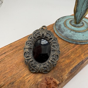 May include: An oval pendant featuring a smooth, solid black center stone encased in an ornate, dark bronze-toned metal frame with intricate scrollwork and a small circular bail at the top for attachment to a chain or cord.