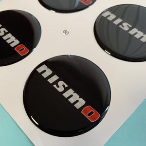 May include: Four black round stickers with the word "NISMO" in silver lettering with a red "O".