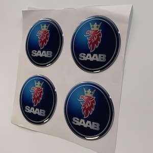 Könnte beinhalten: Vier runde, blaue und silberne Saab-Autoembleme mit einem roten und goldenen Greif-Logo und dem Wort "SAAB" in weißen Buchstaben.