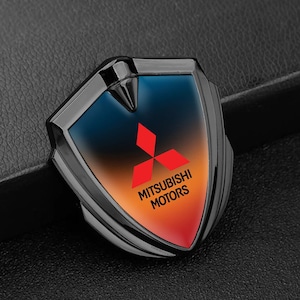 May include: A black and silver Mitsubishi emblem in a shield shape. It has a gradient background of blue, orange, and red, with the Mitsubishi logo and the words "MITSUBISHI MOTORS" in white.