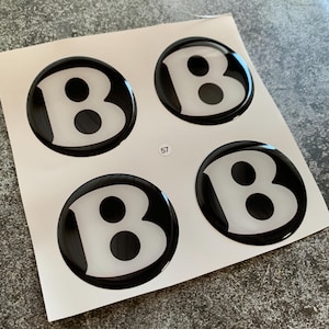 Peut inclure: Quatre autocollants ronds noirs et blancs avec un effet 3D en relief. Chaque autocollant présente une lettre "B" blanche avec deux points noirs à l'intérieur. Les autocollants sont sur une feuille de papier blanche.