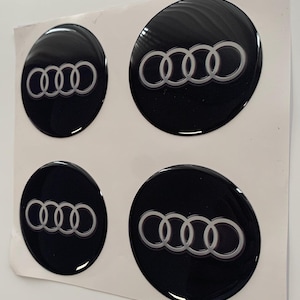 Op de afbeelding: Vier zwarte en zilveren Audi-autoemblemen op een witte achtergrond. De emblemen zijn rond en hebben een glanzende afwerking.