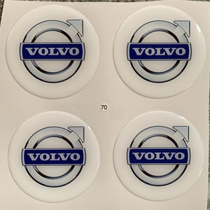 May include: Four white round stickers with a blue Volvo logo. The logo features a silver circle with an arrow pointing to the right and the word "VOLVO" in blue letters.