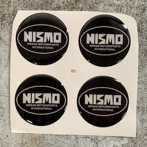 Peut inclure: Quatre autocollants noirs brillants et circulaires avec le texte blanc "NISMO" au-dessus de "NISSAN MOTORSPORTS INTERNATIONAL". Le numéro "60" est imprimé en noir. Les autocollants sont sur un support blanc.