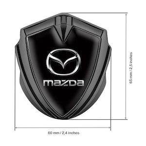 May include: Black shield-shaped emblem with the Mazda logo in silver. The emblem is 2.4 inches wide and 2.5 inches tall. The Mazda logo features a stylized wing design above the brand name.