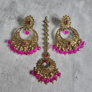 May include: A gold-toned jewelry set featuring a pair of large, ornate earrings and a headpiece. The earrings and headpiece are adorned with pink beads and small white pearls. The set has a traditional design, with a circular shape and intricate detailing.