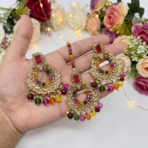 Puede incluir: Juego de joyas doradas con una diadema y pendientes. Cada pieza tiene una gema roja en el centro y está adornada con cuentas de colores en tonos rojos, verdes, amarillos y morados. Los pendientes y la diadema tienen un diseño de filigrana.