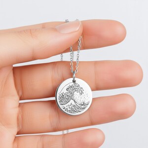 May include: A silver-toned circular pendant necklace featuring a black line art engraving of a large crashing ocean wave. The pendant hangs from a delicate matching silver-toned metal chain, showcasing a detailed artistic design on the surface.