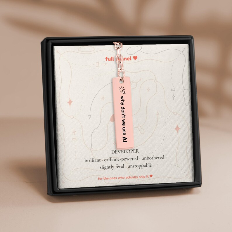 K&ouml;nnte beinhalten: Eine rechteckige Anh&auml;ngerkette in Ros&eacute;gold an einer Kette, pr&auml;sentiert in einer schwarzen Schachtel. Der Anh&auml;nger zeigt ein Cursor-Symbol und den Text "why don't we use AI". Die Karte zeigt "full funnel", "DEVELOPER", "brilliant - caffeine-powered - unbothered - slightly feral - unstoppable" und "for the ones who actually ship it".