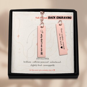 K&ouml;nnte beinhalten: Zwei rechteckige Anh&auml;nger in Ros&eacute;gold an einer Kette, pr&auml;sentiert in einer schwarzen Schachtel. Der linke Anh&auml;nger zeigt ein Cursor-Symbol und den Text "why don't we use AI". Der rechte Anh&auml;nger tr&auml;gt die Aufschrift "Your custom message on the back".