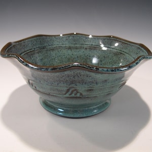 May include: A handcrafted ceramic bowl with a teal glaze and a scalloped rim. The bowl has a pedestal base and a dark brown rim. The exterior features subtle textured details. This decorative bowl is suitable for serving or display.