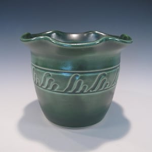 May include: A dark green ceramic pot with a wavy rim and a band of wave-like designs around the middle. The pot has a rounded base and a flared top, and it sits on a white surface.