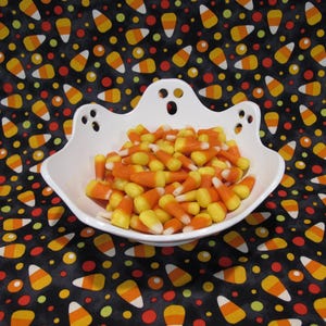 May include: A white ceramic bowl shaped like a ghost with cut-out eyes and a mouth, filled with a pile of orange, yellow, and white candy corn. The bowl features a scalloped rim that mimics the silhouette of a ghost's head and shoulders.