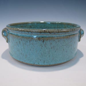 May include: A handmade ceramic bowl with a turquoise glaze and two small handles. The bowl has a speckled texture and a brown rim. The bowl is approximately 3 inches tall and 6 inches in diameter.
