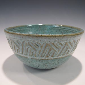 May include: A handmade ceramic bowl with a turquoise glaze and a decorative band of carved geometric patterns. The bowl has a rounded shape and measures approximately 6 inches in diameter and 3 inches in height.