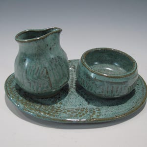 May include: A ceramic set featuring a small pitcher and a bowl resting on an oval tray. The pieces are finished in a speckled teal glaze with visible brown textured patterns and dark brown edges, showcasing a consistent handmade aesthetic.
