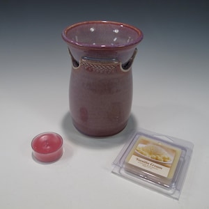 May include: A mauve ceramic wax warmer with a flared rim and textured wheat pattern detail. Beside it sits a small round pink tea light candle and a clear plastic package containing Vanilla Cream scented wax melts with a label.