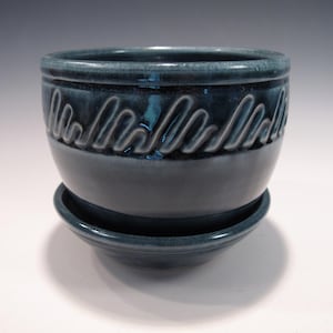 May include: A teal ceramic planter with a matching saucer. The planter has a rounded shape with a textured wave pattern around the top. The glossy finish gives it a handcrafted look, suitable for indoor plants.