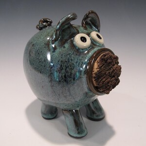 May include: A teal ceramic pig figurine with large, googly eyes and a cork nose. The pig has small ears and a curly tail. The glaze has a speckled appearance. The pig stands on three short legs.