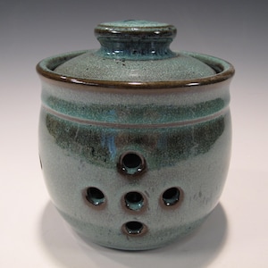 May include: A teal ceramic garlic keeper with a matching lid. The jar has a rounded shape with several circular holes for ventilation. The rim and lid are a darker shade of teal, and the piece has a glossy finish.